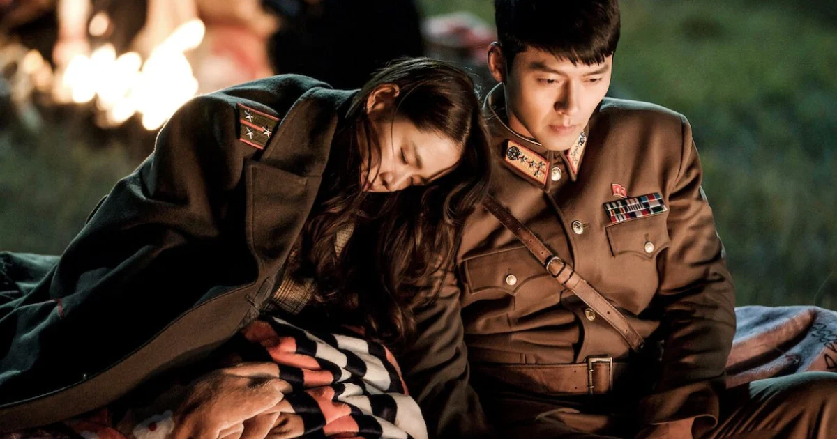 Cena do dorama Pousando no Amor (Crash Landing on You) com Yoon Se-ri dormindo no ombro de Ri Jeong-hyeok ao lado de uma fogueira.